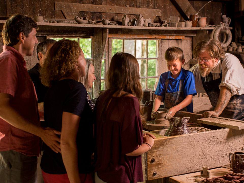 Visitors in the Pottery Shop, a boy works the wheel under the guidance of a costumed historian