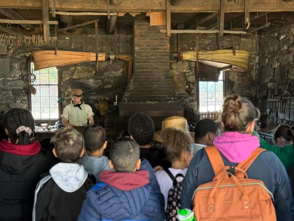 A group of children visit a 19th-century blacksmith shop and chat with a blacksmith