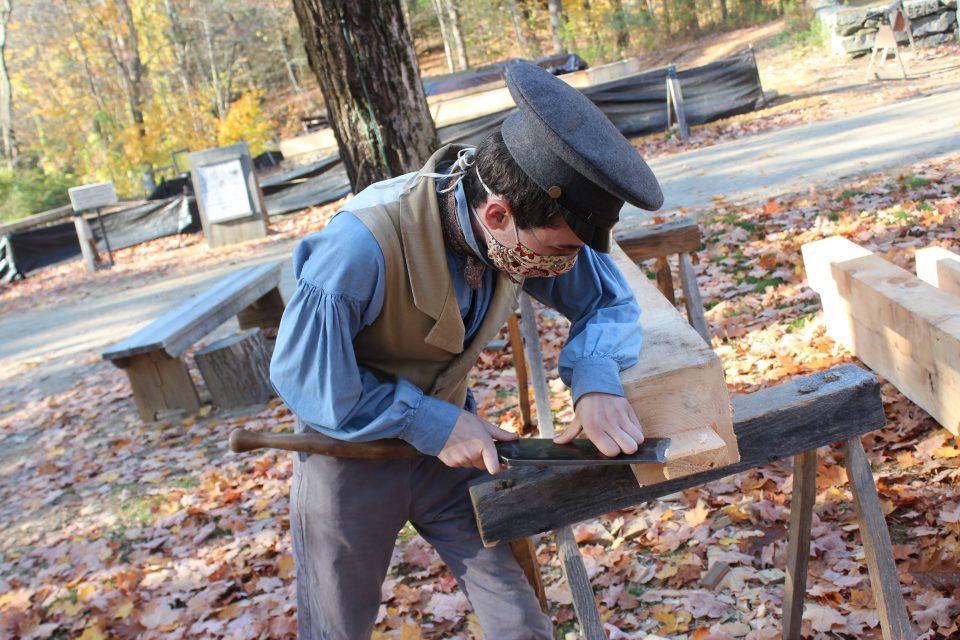 A costumed historian uses a slick on a beam for the frame of the cabinetmaking shop