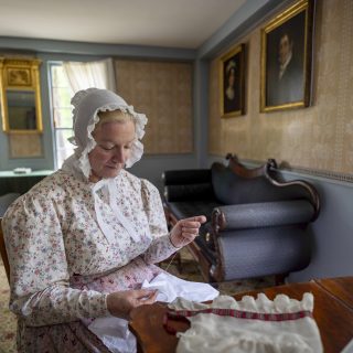 A woman in 1830s dress hand sews a piece of fabric