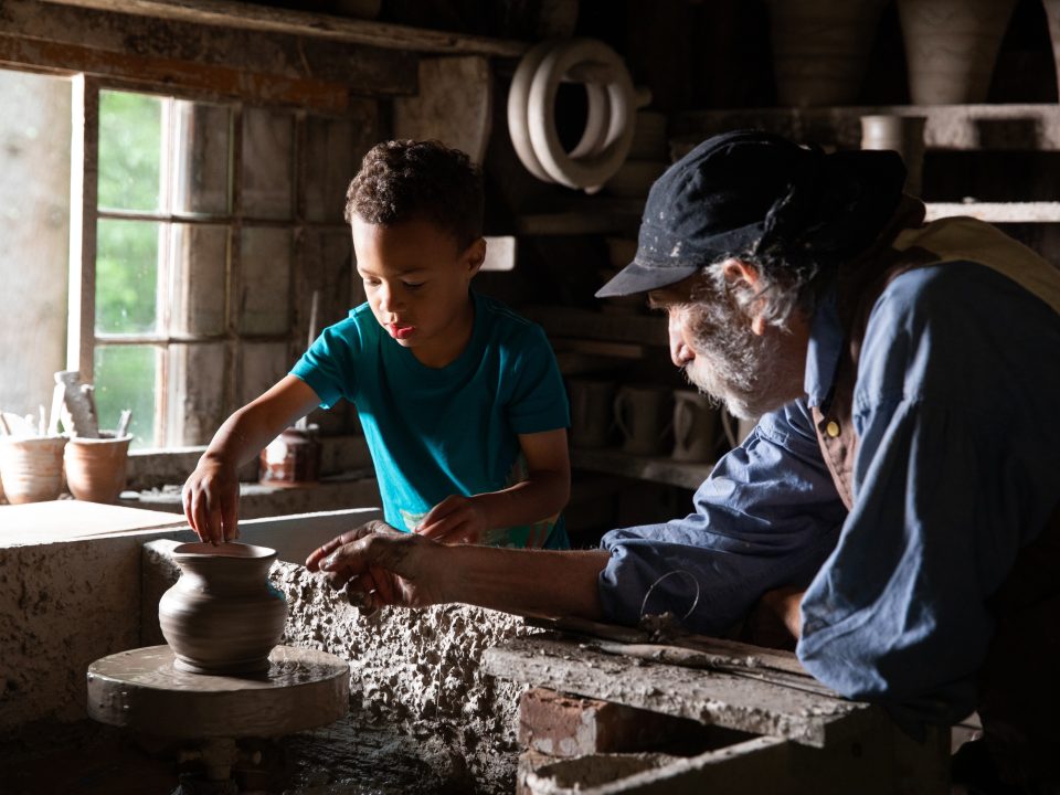 A young child tries his hand at spinning a pot on a wheel, with guidance from a man in 1830s attire.